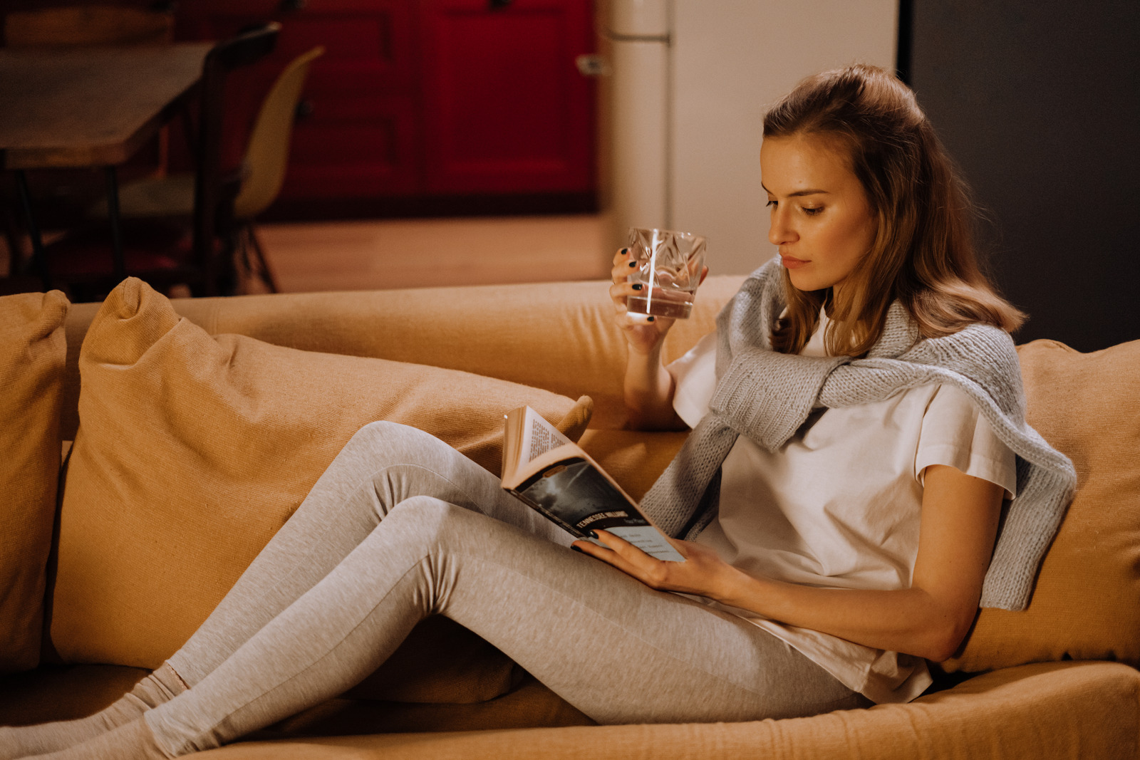 junge Dame sitzt gemütlich auf der Couch, liest ein Buch und trinkt Wein aus einem Weinglas