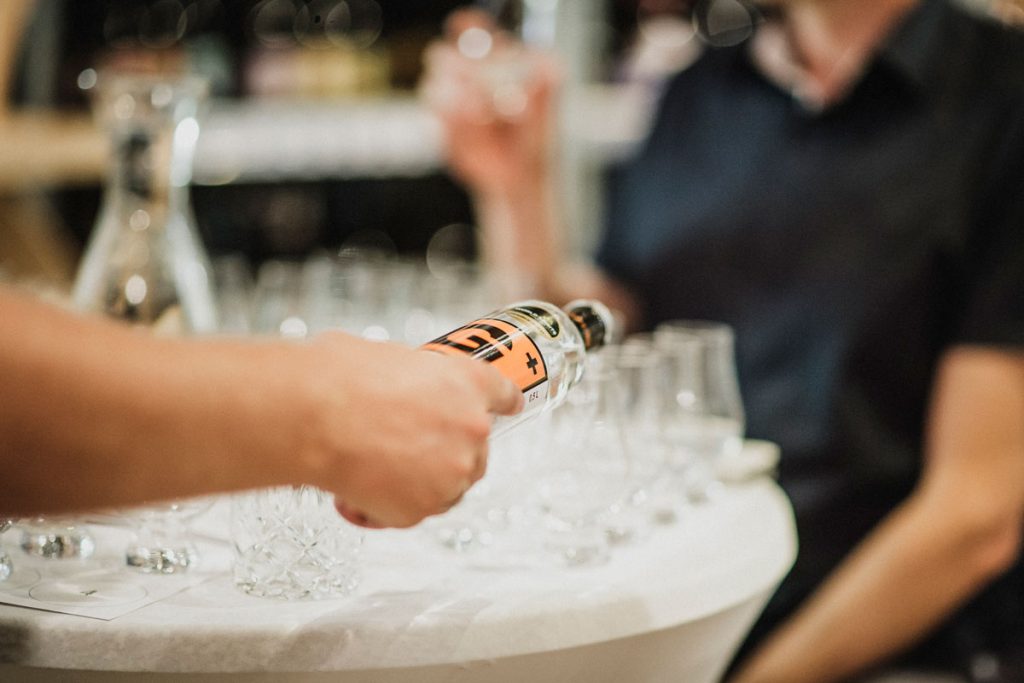 eine Hand, die G+ Gin aus der Flasche in Verkostgläser einschenkt. Im Hintergrund sieht man eine Person in dunklem Shirt.