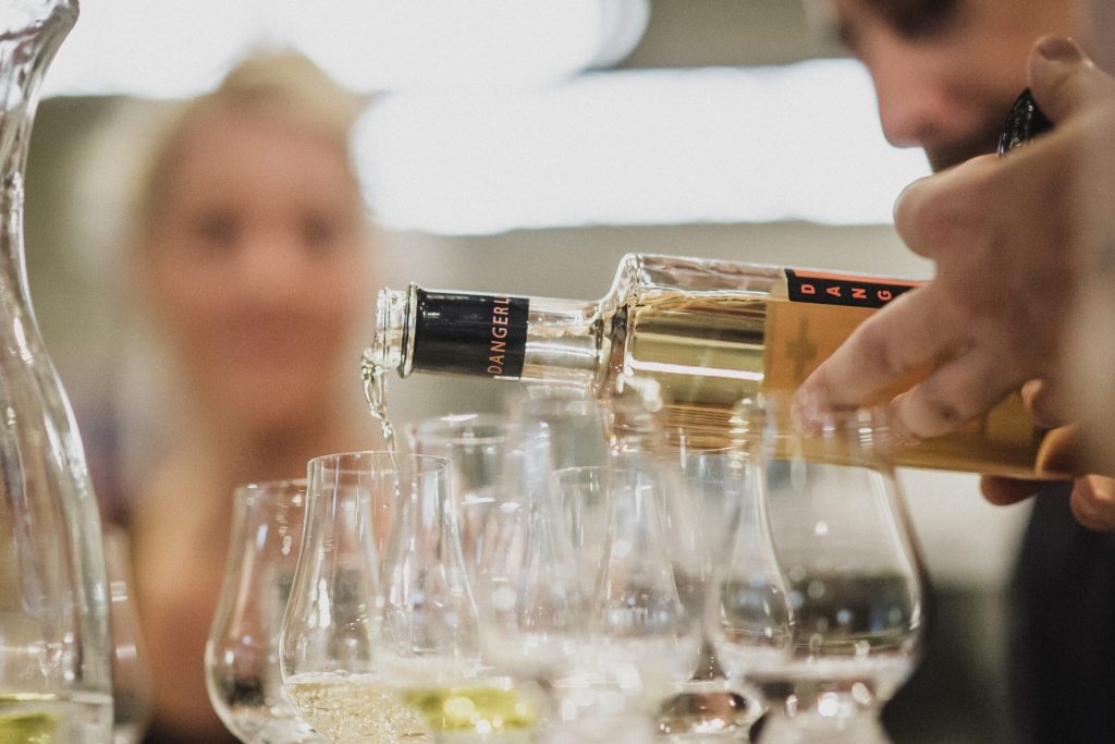 eine Hand, die G+ Gin aus der Flasche in Verkostgläser einschenkt. Im Hintergrund sieht man verschwommen eine blonde Dame sitzen