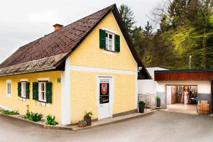 Foto des alten Firmensitzes der Distillery Krauss in Graden. Gelbes Gebäude mit grünen Balken. In der Garage im Nebengebäude sieht man den Brennkessel