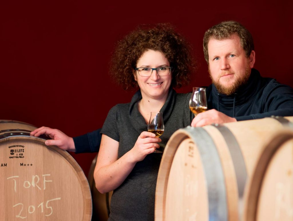 Portraitfoto von Carmen Krauss und Werner Krauss zwischen Whiskyfässern. roter Hintergrund, beide haben ein Verkostglas in der Hand