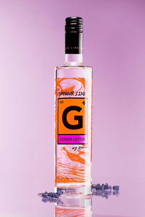 hohe, schlanke 500ml Flasche G+ Flower Edition Gin. Das Etikett spiegelt das Gefahrenzeichen wieder. rosa, lila Hintergrund. Lavendel als Dekoration