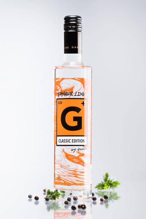hohe, schlanke 500ml Flasche mit G+ Classic Edition Gin. Das Etikett spiegelt das Gefahrenzeichen wider. neutraler Hintergrund. Wacholderbeeren als Dekoration