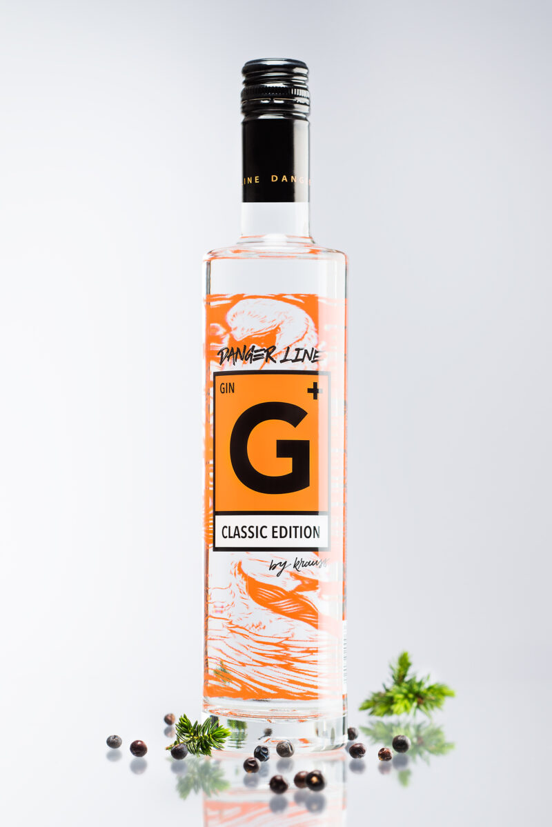 hohe, schlanke 500ml Flasche mit G+ Classic Edition Gin. Das Etikett spiegelt das Gefahrenzeichen wider. neutraler Hintergrund. Wacholderbeeren als Dekoration
