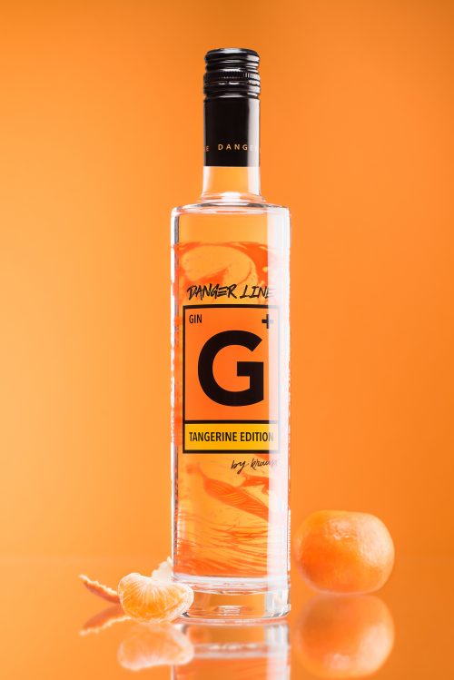 hohe, schlanke 500ml Flasche G+ Tangerine Edition Gin. Das Etikett ist das Gefahrenzeichen. oranger Hintergrund. eine ganze Mandarine und eine Mandarinenspalte als Dekoration