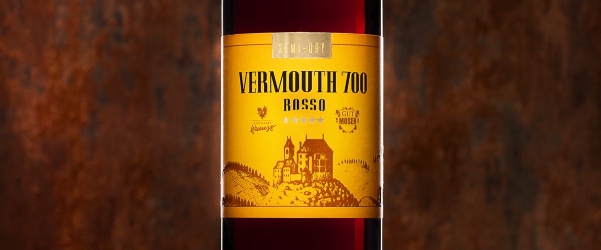 hohe, schlanke 750ml-Flasche gefüllt mit rotem Wermut. Dunkler Hintergrund, Blutorangenscheibe als Dekoration, gelb-oranges Etikett, Vermouth 700 Rosso