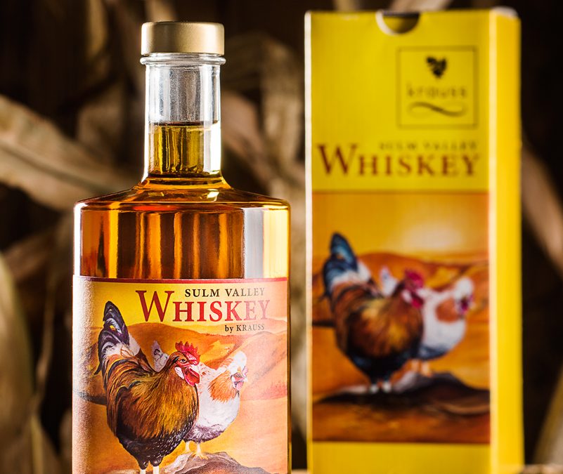 500ml Whisky Flasche mit gelbem Etikett mit zwei Hühnern drauf. gelber Karton im Hintergrund. Erde, Maiskörner und Hühnerfeder als Dekoration. Sulm Valley Whiskey