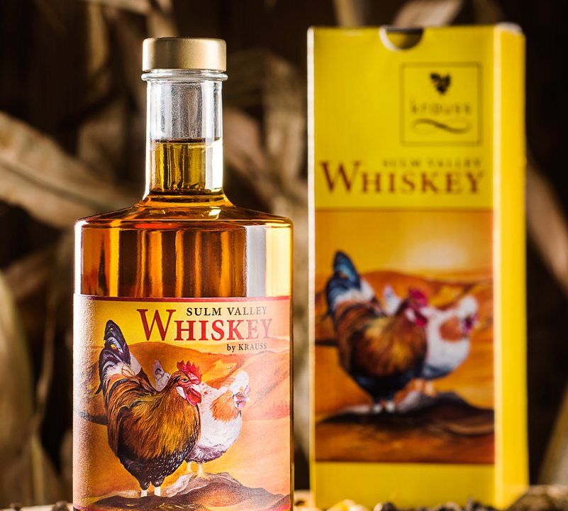 500ml Whisky Flasche mit gelbem Etikett mit zwei Hühnern drauf. gelber Karton im Hintergrund. Erde, Maiskörner und Hühnerfeder als Dekoration. Sulm Valley Whiskey