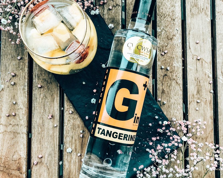 liegende, schlanke G+ Gin Flasche Tangerine mit CWSA Doublegold Medaille, daneben gefülltes Glas mit Eiswürfeln, Blümchen als Dekoration